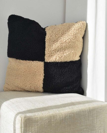 Almohadón decorativo sherpa CHECK by ONYX. Foto en ambiente sobre puff en esquina con sol.